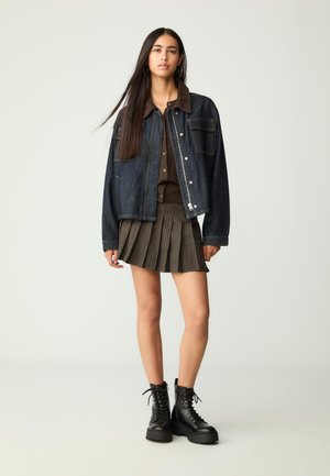 Jeansjacke mit einem braunen Cordkragen, kombiniert mit einem braunen Hemd mit Knöpfen und einem gefalteten dunklen Rock. Schwarze Schnürboots.
