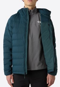 Teal pufferjakke med quiltet mønster, front lynlås og justerbar hætte. Indvendig har den en grå foret skal med lynlås lomme.