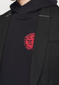 Sudadera negra con un estampado gráfico rojo de una cara sonriente y texto, fabricada en material suave, con un ajuste relajado y un toque de cremallera en el lateral.