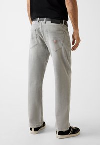 Grijze spijkerbroek met rechte pijpen, vijf zakken en een logopatch aan de achterkant. Draagt zwarte en beige sneakers.