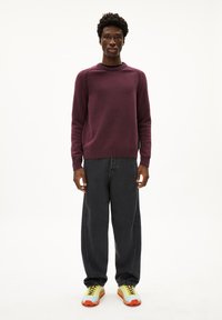 Bordeauxroter Pullover mit geripptem Kragen, kombiniert mit lässigen schwarzen Jeans und mehrfarbigen Sneakers in Gelb-, Blau- und Orangetönen.