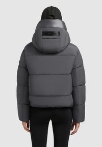 Pelēka pūkaina jaka ar kapuci, ar izšūtu rakstu, augstu apkakli un logo uz muguras. Gluda, spīdīga tekstūra.