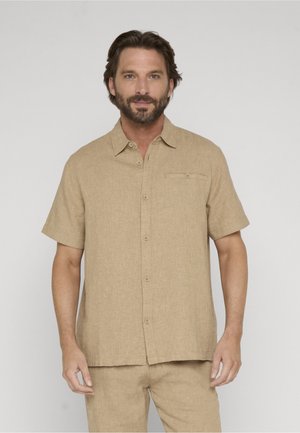 Mężczyzna z brodą, ubrany w beżową koszulę na krótki rękaw z guzikami i pasujące spodnie, stojący na tle jednolitego, jasnego tła.