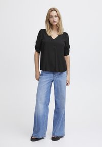 Blusa nera a maniche corte con scollo a V; abbinata a jeans in denim azzurro chiaro a gamba larga; modello indossa sandali neri piatti.