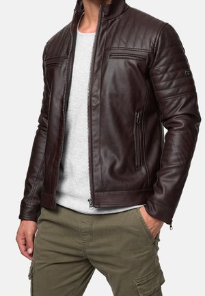 Homme portant une veste matelassée en cuir brun foncé, une chemise gris clair et un pantalon cargo olive, avec une main dans la poche, devant un fond uni.