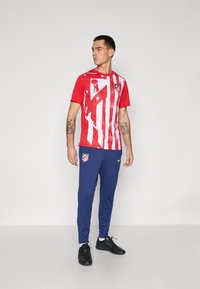 Rød atletisk skjorte med hvide striber og grafik, parret med marineblå svedbukser med logoer. Sorte fodtøj fuldender looket.