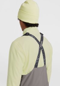 Grijze overalls met zwarte O'Neill schouderbanden, gecombineerd met een felgele gebreide beanie, tegen een eenvoudige achtergrond. Zachte textuur en functioneel ontwerp.