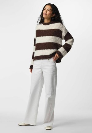Femme aux cheveux bouclés portant un pull rayé marron et blanc, un pantalon blanc à jambes larges et des chaussures plates couleur crème, debout avec une main sur la hanche.
