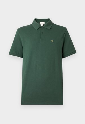 Ciemnozielona koszulka polo z krótkim rękawem, z dwoma guzikami i małym żółtym logo "cK" na lewej klatce piersiowej, na jednolitym tle.
