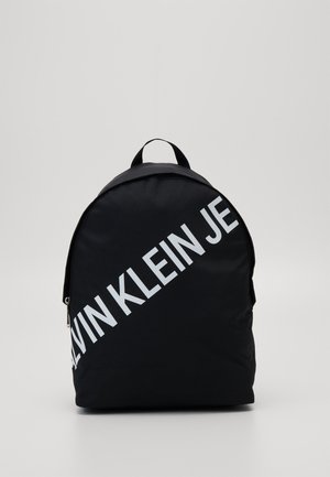 Sort rygsæk med en diagonal hvid "CALVIN KLEIN JE"-logo på fronten, udstyret med et topgreb og lynlåslukning.