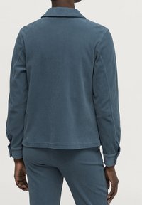 Modrá kordurová bunda s límcem a dlouhými rukávy, s rovným střihem a jemnou texturou; zadní pohled ukazuje detaily švů.