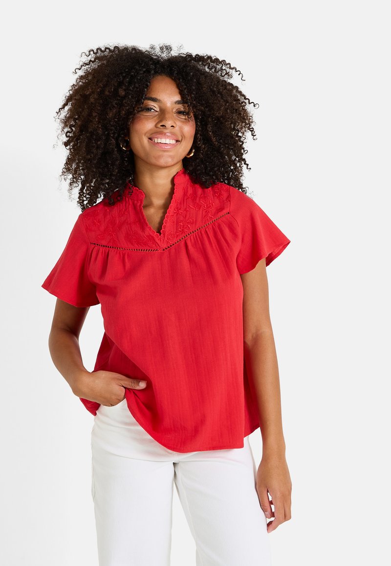 Blouse rouge à manches courtes avec un col haut, ornée de dentelle et d'une coupe ample. Le tissu semble lisse et léger.
