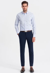 Camicia azzurro chiaro con piccoli pois, pantaloni navy su misura, cintura marrone e scarpe scure. Tessuto liscio, design aderente.