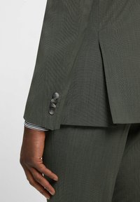 Dark green suit jacket fabric with a textured finish. Four gray buttons on the sleeve. White and black striped shirt cuff visible.