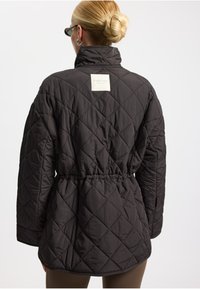 Zwarte gewatteerde jas met hoge kraag, getailleerde taille en een label op de achterkant. De stof heeft een gladde, matte textuur en een ruitpatroon.