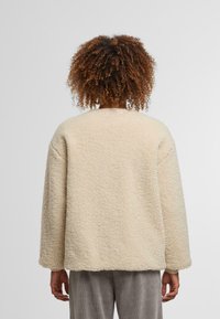 Kremowa, pulowerowa kurtka o miękkiej, puszystej fakturze, luźnym kroju i opuszczonych ramionach. Okrągły dekolt i długie rękawy, bez widocznych zapięć.