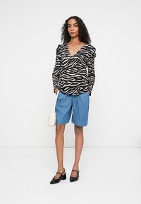 Top a maniche lunghe a strisce zebrate nere e crema con spalle gonfie, abbinato a pantaloncini di denim blu e décolleté slingback neri, mentre tiene una borsa bianca.