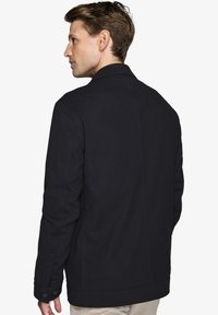 Sort jakke med en struktureret design, der har en glat tekstur, knappede manchetter og en klassisk krave. Bagudsigten viser rene linjer.