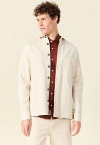 Lichtcrème overhemd met knopen, voorzien van twee borstzakken, zwarte knopen en een losse pasvorm over een donkerrood overhemd eronder.