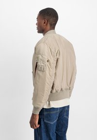 Beige bomber jacket with ribbed cuffs and hem, made of a smooth material. Features a utility pocket on the sleeve and a zipper closure.