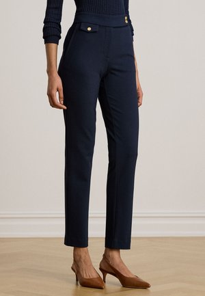 Femme portant un pantalon slim bleu marine avec des détails de boutons dorés, associé à des talons pointus marron et un pull côtelé bleu marine.
