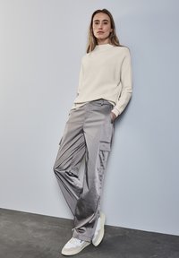 Pantaloni cargo grigi chiari con tasche laterali, abbinati a un maglione crema a collo alto e sneaker bianche su un pavimento grigio contro una parete chiara.