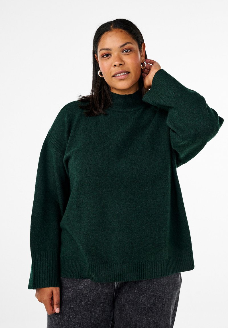 Temno zelena, pletena turtleneck majica z udobnim krojem in rebrastimi zapestji. Model se pozira z eno roko blizu ušesa, kar prikazuje oblačilo.
