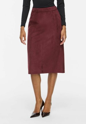 Femme portant une jupe crayon bordeaux longueur genoux associée à un haut noir à manches longues et des chaussures à talons aiguilles noires pointues.