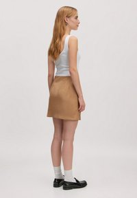 Brauner Satinsrock mit ausgestellt Design, kombiniert mit einem weißen Tanktop. Das Modell trägt schwarze Loafers und graue Socken, seitlich gezeigt.