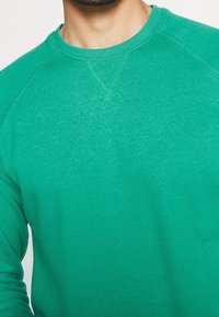 Sudadera verde con cuello redondo, mangas raglán y tejido texturizado. Presenta costuras visibles y un acabado liso y suave.