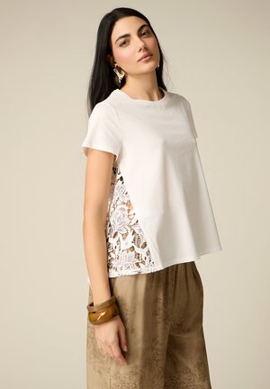 Donna con lunghi capelli scuri che indossa una maglietta bianca a maniche corte con pannelli laterali in pizzo, pantaloni marroni con fantasia, grandi orecchini dorati e due braccialetti marroni.