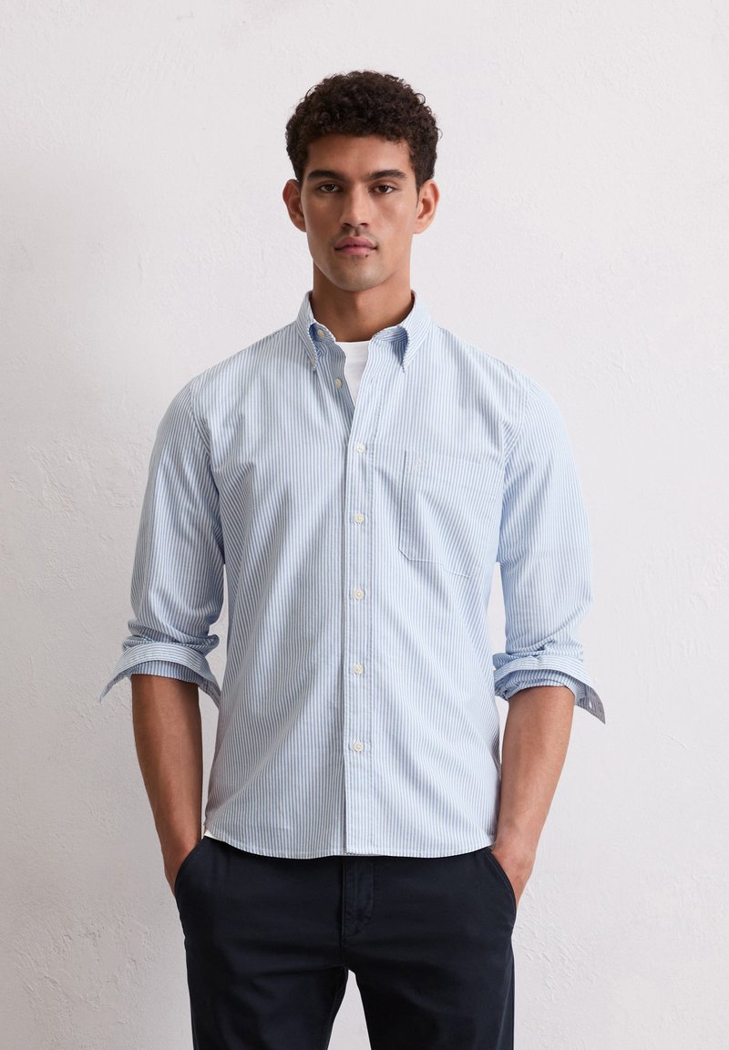 Camisa de button-down a rayas azul claro y blanca con cuello, un bolsillo en el pecho, mangas largas remangadas, de tela de algodón, combinada con pantalones oscuros.