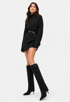 Vrouw draagt een zwarte coltrui sweaterjurk met een riem en kniehoge zwarte suède haklaarzen tegen een effen achtergrond.