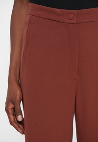 Gros plan sur la main d'une personne à côté de la taille d'un pantalon couleur rouille avec un bouton recouvert de tissu assorti et des poches latérales.