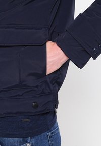 Main dans la poche d'une veste bleu marine avec poche à rabat et poignet boutonné, portée par-dessus une chemise foncée et un jean bleu.