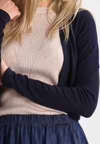 Marineblauer Strickcardigan mit langen Ärmeln, geschichtet über einem hellen gerippten Pullover, kombiniert mit einem Marineblauem Rock mit elastischem Bund. Weiche Textur, lässige Passform.