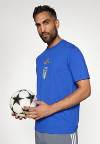 Μπλε μπλουζάκι με στρογγυλή λαιμόκοψη και χρυσές λεπτομέρειες, με λογότυπο Ιταλίας, φορεμένο από έναν άνδρα που κρατάει μια άσπρη και μαύρη ποδοσφαιρική μπάλα με σχέδια άστρων.