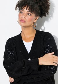 Černý pletený kardigan se vzory v krajkovém stylu, V-čelním výstřihem a jedním knoflíkem, spárovaný s bílým tričkem pod ním.