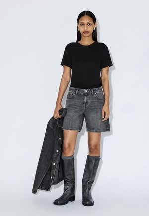 T-shirt noir à manches courtes, shorts en denim noir délavé, et bottes grises montantes jusqu'aux genoux. Le mannequin tient une veste en denim noire.