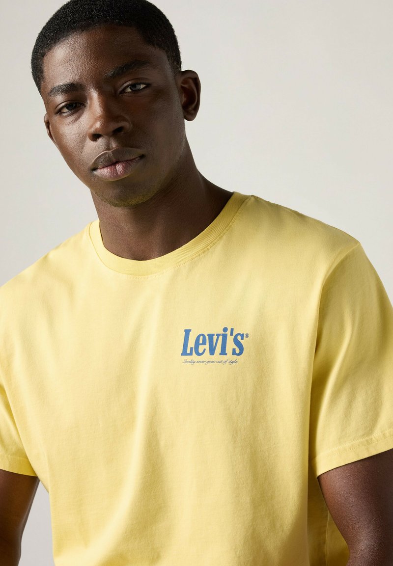 Junger schwarzer Mann trägt ein hellgelbes Levi's T-Shirt mit blauem Logo auf der Brust, schaut nach vorne vor einem neutralen Hintergrund.