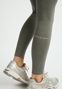 Leggings da allenamento verde oliva con un sottile motivo geometrico, con dettaglio del logo sulla gamba inferiore. Abbinati a scarpe sportive grigie e argentate.