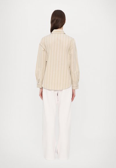 Beige striped shirt with a relaxed fit, long sleeves, and a slightly curved hem, paired with white wide-leg trousers.