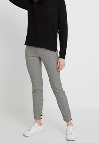Sudadera negra con un cuello alto, combinada con pantalones ajustados de cuadros en blanco y negro. Zapatillas blancas completan el look.