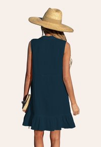 Abito senza maniche blu navy con orlo a volants, realizzato in tessuto testurizzato. Il modello indossa un cappello di paglia e tiene una borsa a mano in tessuto naturale.