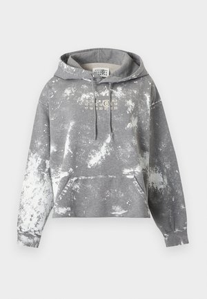 Μαύρο άτομο φορά γκρι φούτερ με tie-dye, ανοιχτό μπλε ξεβαμμένα τζιν και μαύρα μποτάκια μέχρι τον αστράγαλο, στέκεται μπροστά σε απλό ανοιχτό φόντο.