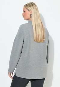 Maglia grigia a maniche lunghe realizzata in materiale morbido, con colletto alto, spacco laterale e vestibilità ampia, vista da dietro.