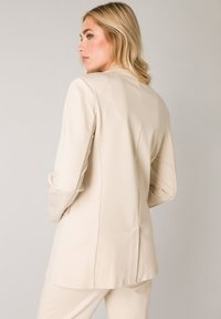Beige op maat gemaakte blazer met een gestructureerd ontwerp, lange mouwen en een gladde textuur. De achterkant heeft een enkele naad en een lichte ventilatie.