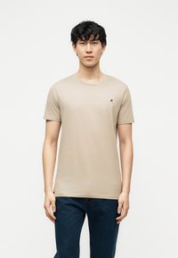 Νεαρός άνδρας με μαύρα σγουρά μαλλιά, φορώντας μπεζ κοντομάνικο μπλουζάκι και σκούρο μπλε τζιν, στέκεται μπροστά σε απλό λευκό φόντο.