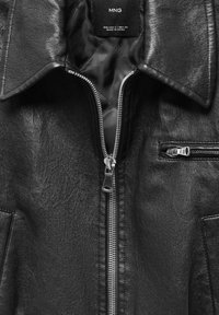 Veste en cuir noire avec fermeture éclair argentée partiellement ouverte, col avant, et une petite poche zippée du côté droit, étiquetée MNG, fabriquée en Chine.