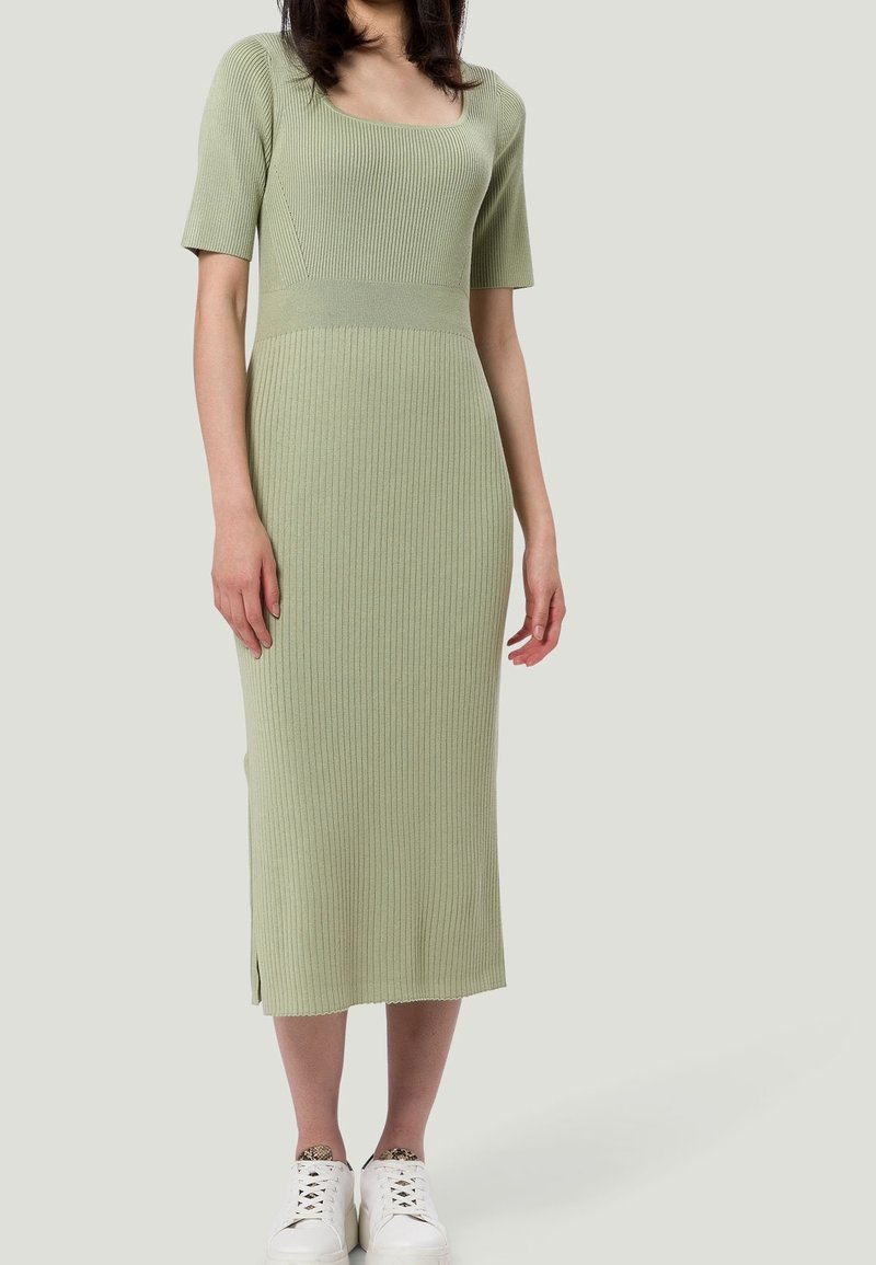 Femme portant une robe midi côtelée vert clair avec des manches courtes et un col carré, associée à des baskets blanches.